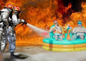 Disaster first responders spraying water on fools in a inflatable pool, surrounded by raging fire.