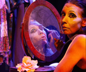 Föreställningsfoto från Kraftverksprinsessan. En sminkad kvinna tittar mot kameran, medan hennes spegelbild tittar på sig själv och sminkar sig.