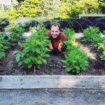 Tre personer som ligger och ler mot kameran mellan odlingsraderna i en matträdgård.