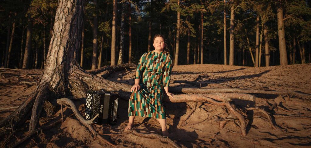 Ett fribas-drapspel står vid roten av en tall. På en av rötterna sitter en barfota musiker i grön klänning, Larisa Ljungkrona-Mönttinen.