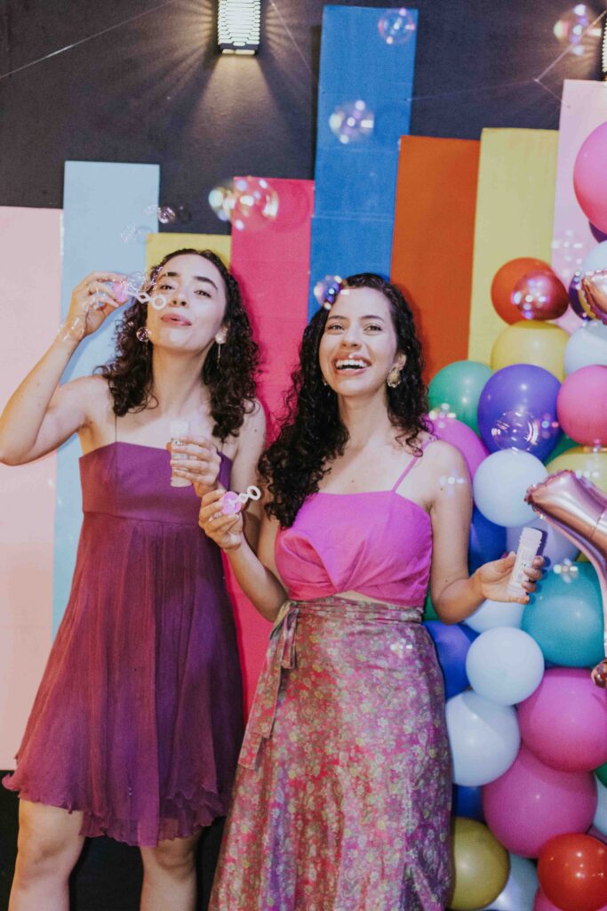 Två glada kvinnor i festklänningar står framför en vägg full av färger och ballonger och blåser såpbubblor