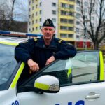 Kommunpolis vid sin polisbil, höga bostadshus i bakgrunden